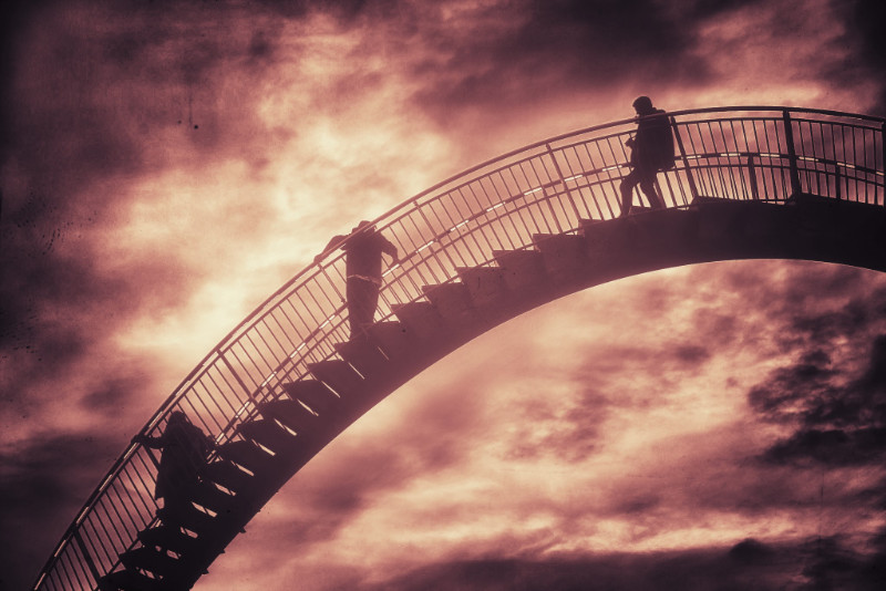 Three people are standing on what looks like a mix between a bridge and a stairway. The construction enters the frame on the right and slopes towards the bottom left edge of the frame, with no supporting structure visible. The people are facing left and looking towards the background, which consists of a cloudy sky in ominous red and brown colors.