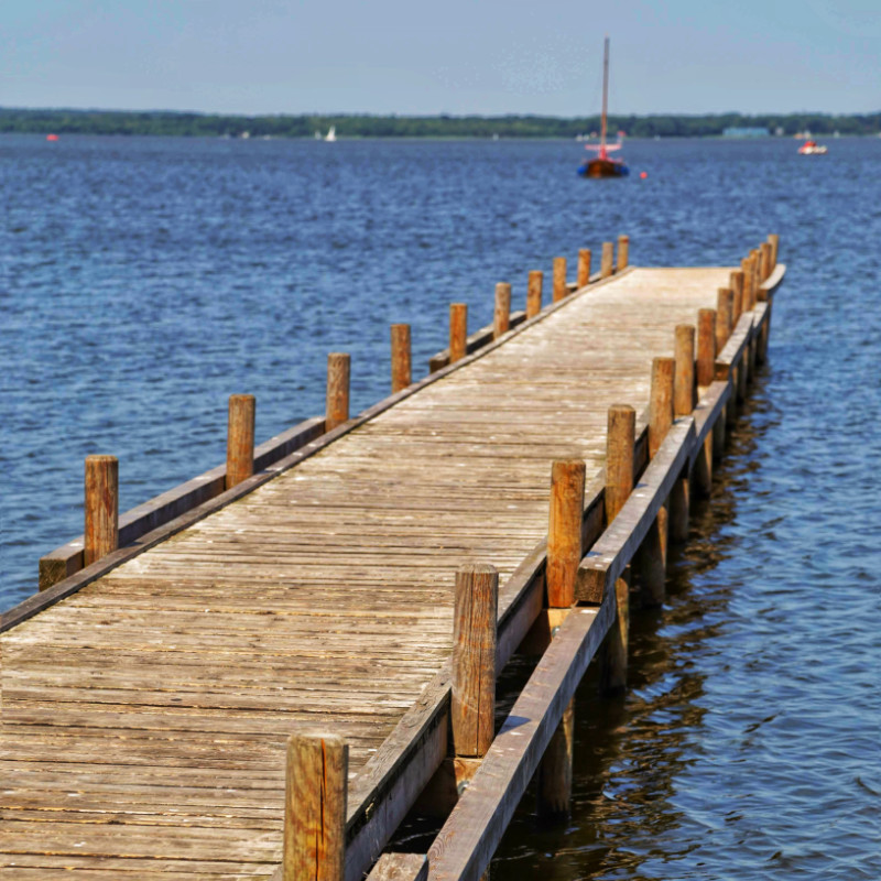 Depicted is a wooden pier, with the view out towards the water. The pier is empty, but a single boat is approaching. In the background, sails of other ships and the distant shore are visible.